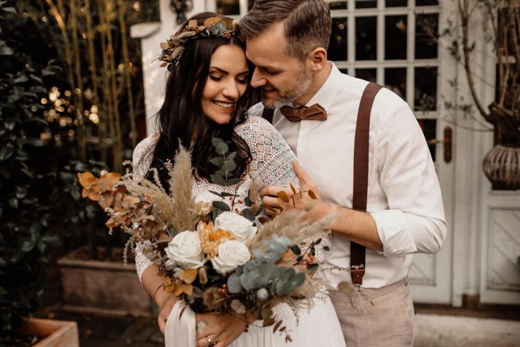 Boho-Brautpaar vor einer weißen Sprossentür, Braut mit offenen dunklen Haaren, Blumenkranz und Pampasgras-Strauß