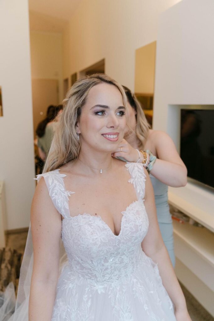 Lächelnde Braut mit offenen blonden Haaren, beim Anziehen wird ihr die Kette angelegt