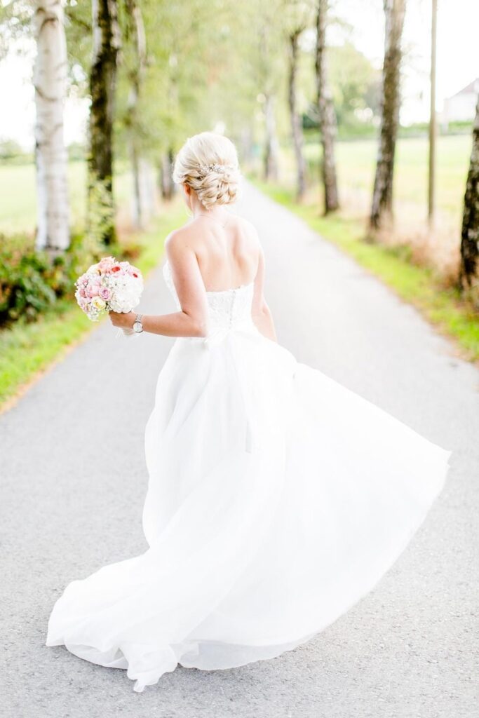 Braut mit blonder Hochsteckfrisur und Brautstrauß läuft im schwingenden Kleid eine Birkenallee entlang