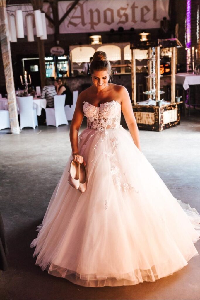 Braut mit Dutt im Ballkleid hält auf der Hochzeitsfeier ihre Schuhe in der Hand
