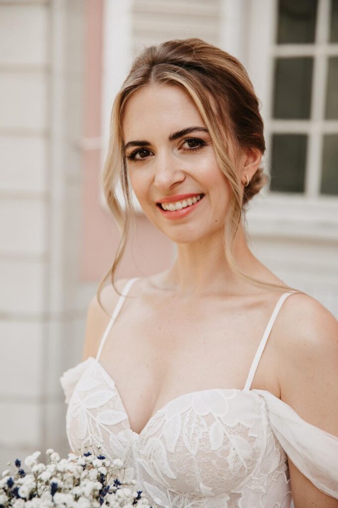 Lächelnde Braut mit natürlichem Braut-Make-up, korallfarbenen Lippen und lockerer Hochsteckfrisur im Spitzenkleid