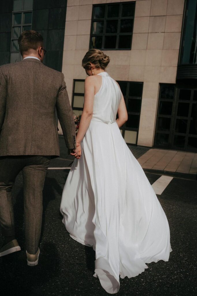 Brautpaar geht Hand in Hand, Braut mit eleganter Hochsteckfrisur im fließenden Neckholder-Kleid
