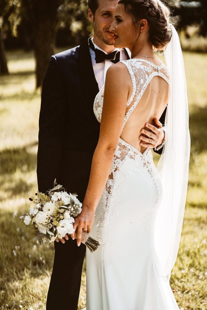 Braut mit Hochsteckfrisur und Schleier im Spitzenkleid mit Rückenausschnitt an der Seite des Bräutigams