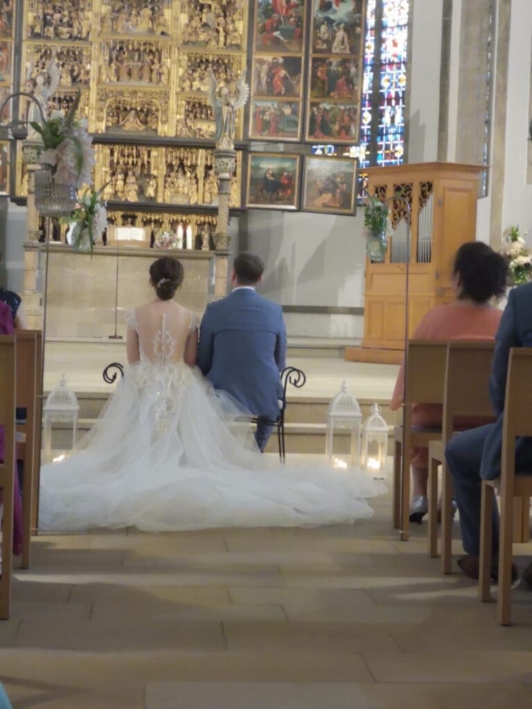 Brautpaar sitzt bei der kirchlichen Trauung vor dem Altar, Braut mit Dutt und Kleid mit langer Schleppe