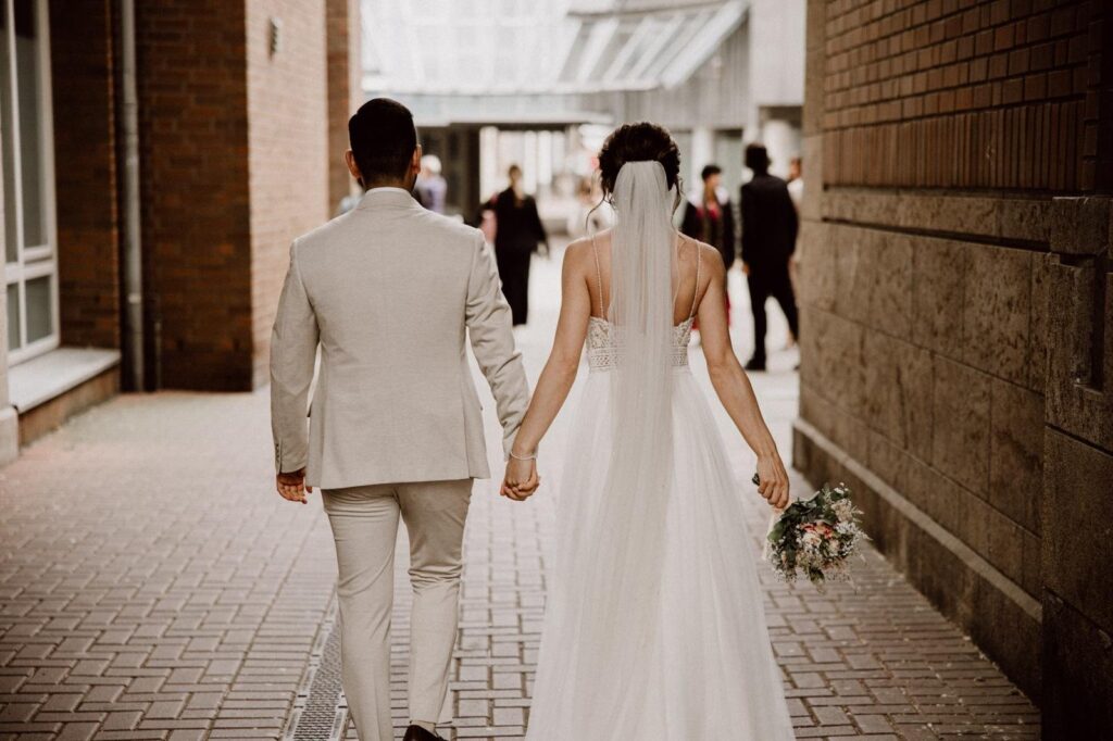 Brautpaar Hand in Hand von hinten, Braut mit Hochsteckfrisur, langem Schleier und Spitzenkleid