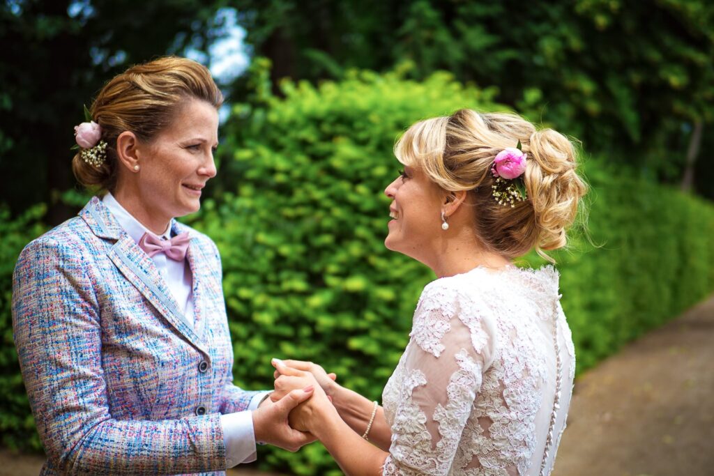 Zwei Bräute mit Hochsteckfrisuren und Blüten im Haar halten sich bei ihrer Hochzeit an den Händen