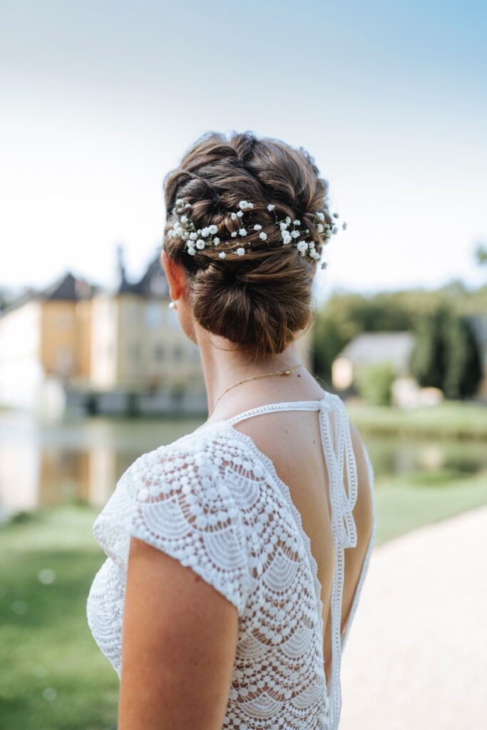 Geflochtene Hochsteckfrisur mit Schleierkraut, Braut im Spitzenkleid vor einem Schloss am Wasser