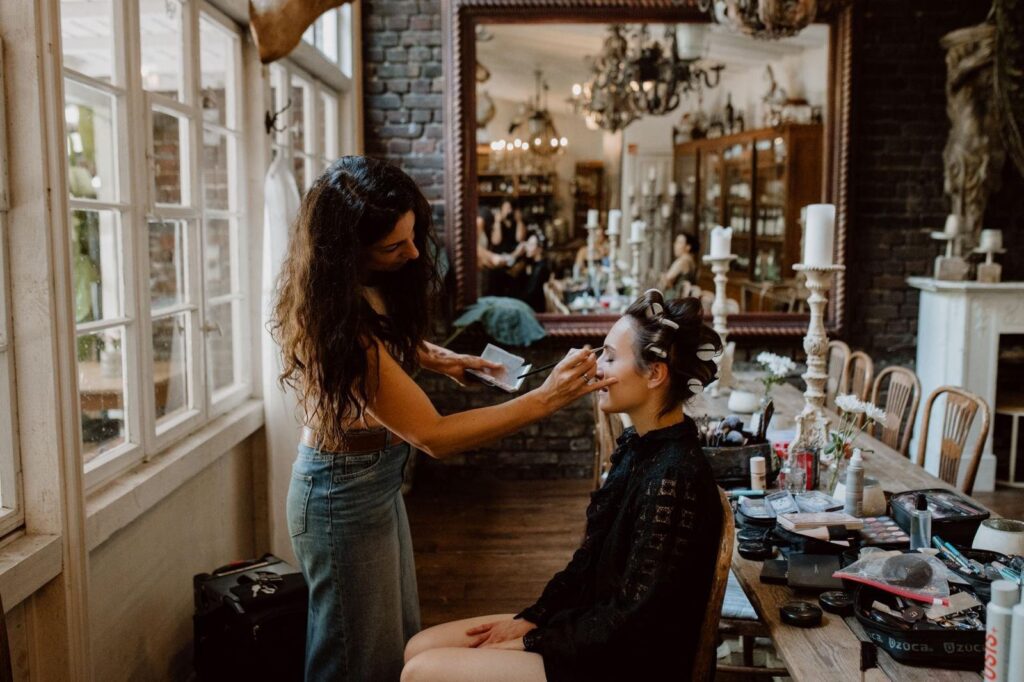 Natalie Aust schminkt die Braut mit Lockenwicklern an einem langen Tisch voller Make-up in einer Vintage-Location