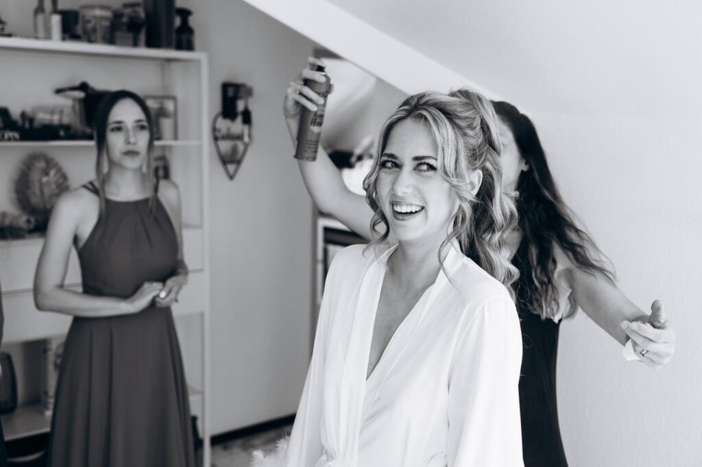 Lachende Braut im Morgenmantel, Natalie Aust fixiert die Locken mit Haarspray, eine Brautjungfer schaut zu