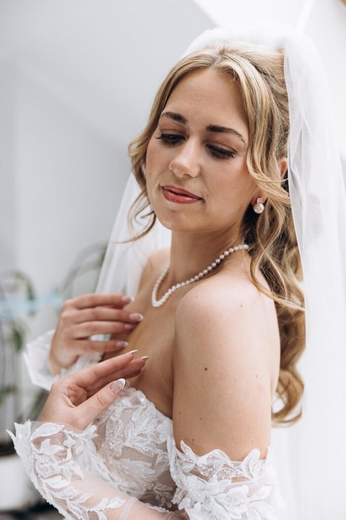 Braut mit halboffener Frisur aus weichen Wellen, Schleier, Perlenkette und schulterfreiem Spitzenkleid