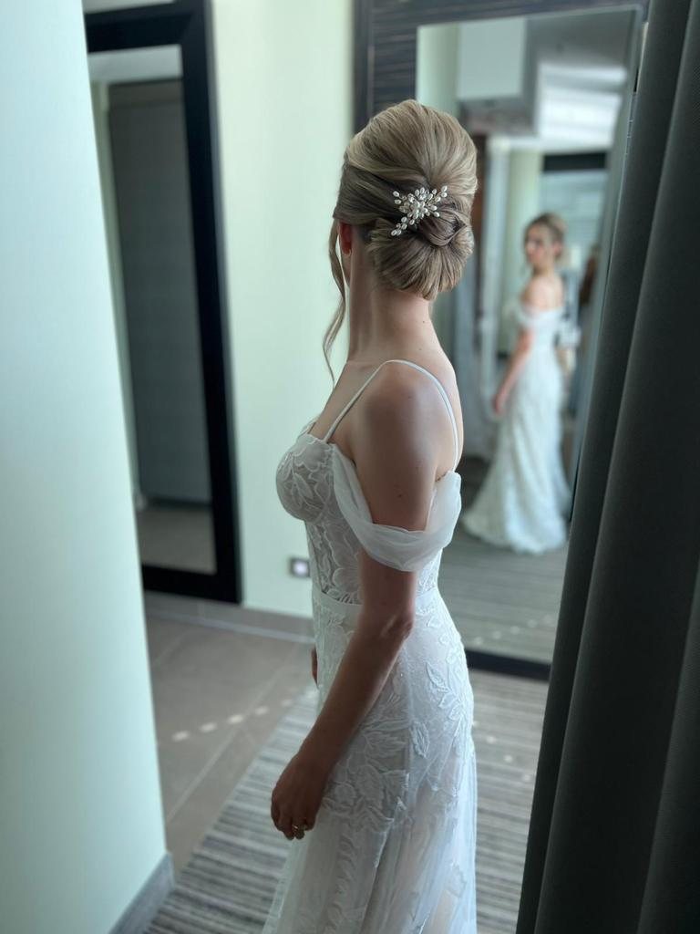 Braut mit glattem, hoch gestecktem Knoten und Haarschmuck vor dem Spiegel im schulterfreien Kleid