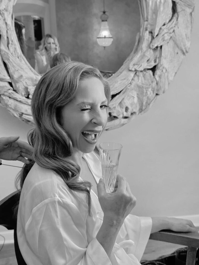 Lachende Braut mit Hollywood Waves zwinkert beim Getting Ready mit einem Glas Sekt in die Kamera