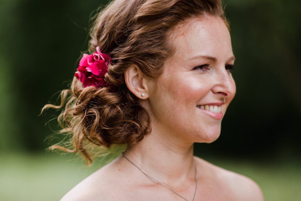 Lachende Braut mit seitlicher Lockenhochsteckfrisur und pinker Blüte im Haar